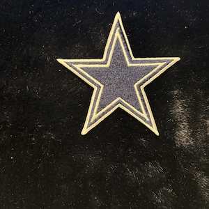 Puede incluir: Un parche en forma de estrella azul marino con un borde blanco. El parche es probablemente para ropa o accesorios, con un diseño de estrella clásico. La estrella está centrada en un fondo oscuro y texturizado.