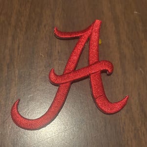 May include: A red embroidered patch in the shape of a stylized capital letter A. The embroidery features a dense, textured thread pattern with a slight sheen, forming the classic script-style letter with curved terminals at the top and bottom.