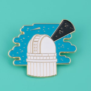 Könnte beinhalten: Goldfarbene Emaille-Anstecknadel mit einem weißen Observatoriumsgebäude, auf dem ein schwarzes Teleskop auf ein Sternenfeld vor blauem Himmel gerichtet ist.