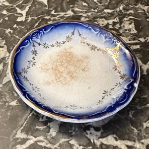 May include: A small, white ceramic saucer with a blue and gold rim. The saucer has a delicate floral pattern in gold and is slightly chipped.