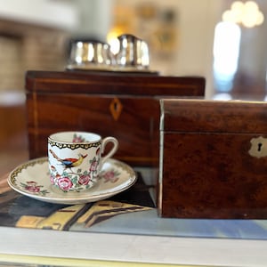 May include: A white teacup and saucer with a floral and bird design sits on top of a stack of books. Two wooden boxes with intricate inlays are in the background.