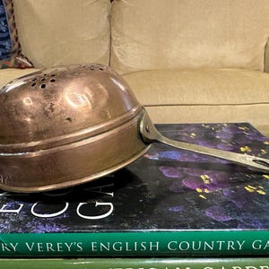 May include: A vintage copper colander with a handle. The colander has a hammered finish and is sitting on top of a stack of books.