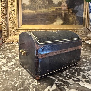 Puede incluir: Peque&ntilde;a caja antigua de cuero negro con tapa curvada y ribete dorado. La caja tiene un cierre de lat&oacute;n y probablemente se utiliza para joyas o peque&ntilde;os recuerdos. Colocada sobre una superficie de m&aacute;rmol.
