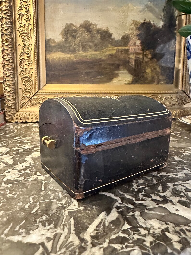 Puede incluir: Peque&ntilde;a caja antigua de cuero negro con tapa curvada y ribete dorado. La caja tiene un cierre de lat&oacute;n y probablemente se utiliza para joyas o peque&ntilde;os recuerdos. Colocada sobre una superficie de m&aacute;rmol.