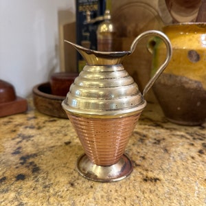 May include: A small, copper and brass pitcher with a fluted design. The pitcher has a round base and a handle.