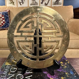 May include: A gold-colored metal decorative wall hanging with a circular design featuring a traditional Chinese symbol. The wall hanging is on a black stand and is resting on a book.
