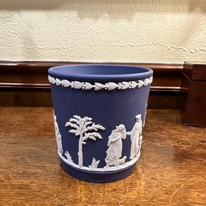 May include: A blue and white ceramic planter with a raised design of people and a tree. The planter is made in the style of Wedgwood.