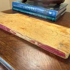 May include: A rectangular wooden cutting board with a handle, featuring a natural light wood grain surface and worn red paint along the edges. The board shows signs of use with visible scratches and texture across the top surface.