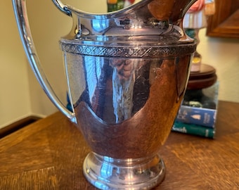 May include: A silver-plated pitcher with a curved spout and a handle. The pitcher has a decorative band around the top and sits on a round base. The pitcher is reflecting the surrounding environment.