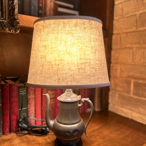 May include: A vintage pewter teapot lamp with a beige linen lampshade. The lamp is on a wooden table with books in the background.