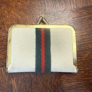 May include: A rectangular coin purse made of off-white canvas fabric featuring a vertical central stripe in dark green and red. The purse is framed with a gold-toned metal border and includes a top-mounted metal kiss-lock clasp closure.
