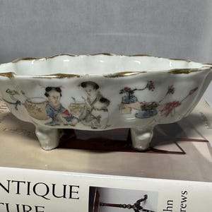 May include: A white porcelain bowl with three legs and a gold rim. The bowl is decorated with hand-painted figures and floral designs in shades of pink, blue, and green.