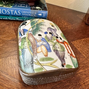 May include: A rectangular porcelain box with a silver rim and a decorative design of five children in a garden setting. The children are wearing colorful clothing and are surrounded by green trees and yellow flowers. The box is on a wooden surface.
