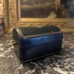 Puede incluir: Una peque&ntilde;a caja de joyer&iacute;a antigua con una parte superior curvada. La caja es azul marino con ribetes dorados y un cierre de lat&oacute;n. La parte superior est&aacute; cubierta de terciopelo negro. La caja est&aacute; sobre una superficie de m&aacute;rmol.