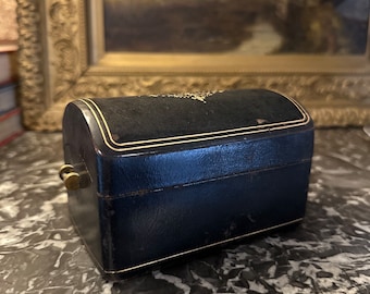 May include: A small, antique jewelry box with a curved top. The box is navy blue with gold trim and a brass clasp. The top is covered in black velvet. The box is sitting on a marble surface.