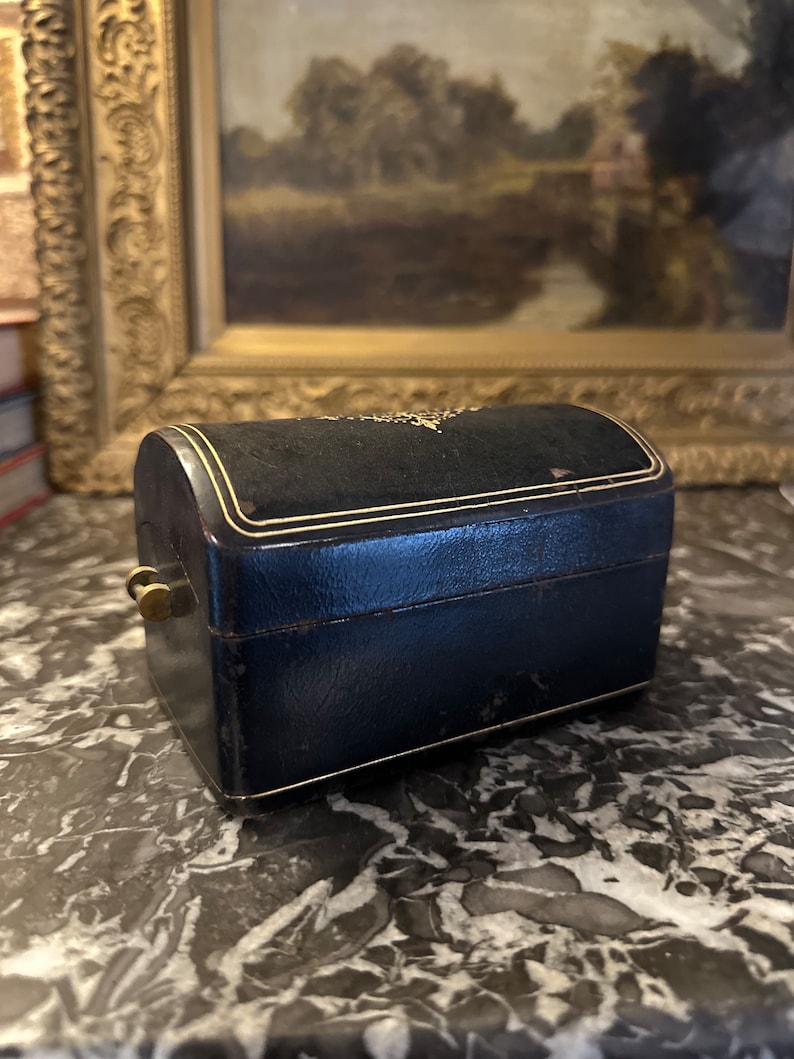 Puede incluir: Una peque&ntilde;a caja de joyer&iacute;a antigua con una parte superior curvada. La caja es azul marino con ribetes dorados y un cierre de lat&oacute;n. La parte superior est&aacute; cubierta de terciopelo negro. La caja est&aacute; sobre una superficie de m&aacute;rmol.