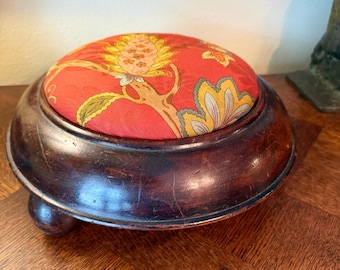 May include: A circular wooden pincushion with a dark brown polished finish and a rounded base. The top features a padded fabric insert in a vibrant red color, decorated with a detailed botanical pattern in shades of yellow, green, and orange.