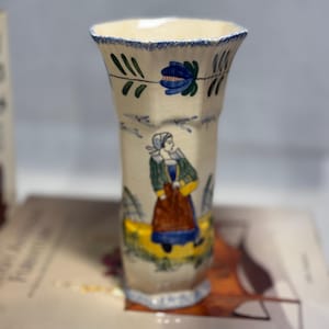 May include: A white ceramic vase with a blue and green floral design and a painted image of a woman in a yellow dress. The vase has a hexagonal shape and a flared top.