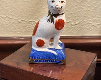 May include: A white ceramic cat figurine with orange spots and a brown bow sitting on a blue and gold cushion.