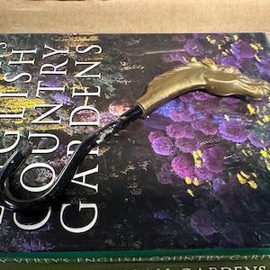 May include: A gold-colored horse head cane handle with a black metal shaft and a hook at the end. The cane is resting on a book titled "Rosemary Verey's English Country Gardens".