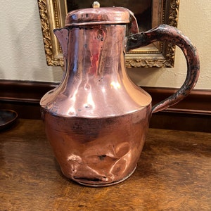 May include: A copper pitcher with a rounded body and a handle. The pitcher has a lid with a small knob on top. The pitcher is sitting on a wooden surface.
