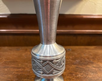 May include: A silver pewter vase with an ornate design around the middle. The vase has a flared top and a round base.