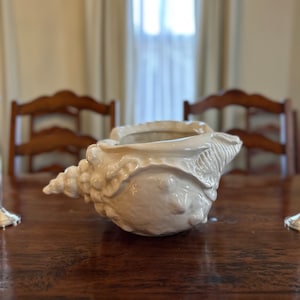 May include: A cream-colored, ceramic seashell-shaped decorative bowl. The bowl is placed on a dark wooden table. The seashell has a textured surface with detailed ridges and a pointed spiral end. The background includes a window and wooden chairs.