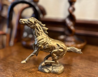 May include: A gold-colored metal figurine of a horse rearing up on its hind legs. The horse is detailed with a flowing mane and tail. The figurine is on a wooden surface.