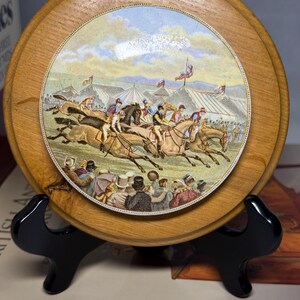 May include: A round, wooden decorative plate featuring a detailed illustration of a horse race. The illustration depicts jockeys on horseback, spectators, and flags under a cloudy sky. The plate is displayed on a black stand.