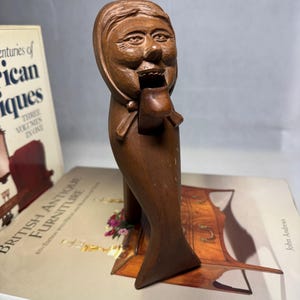 May include: A carved wooden nutcracker in the shape of a woman with a bonnet. The nutcracker is brown and has a detailed face. The nutcracker is standing on a book titled "British Antique Furniture".