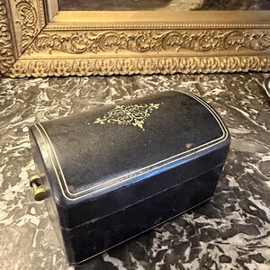 Puede incluir: Una caja de joyer&iacute;a negra, rectangular, con la parte superior curvada. La caja tiene ribetes dorados y un dise&ntilde;o decorativo dorado en la tapa. Se ve un cierre dorado en el lateral. La caja est&aacute; sobre una superficie de m&aacute;rmol.