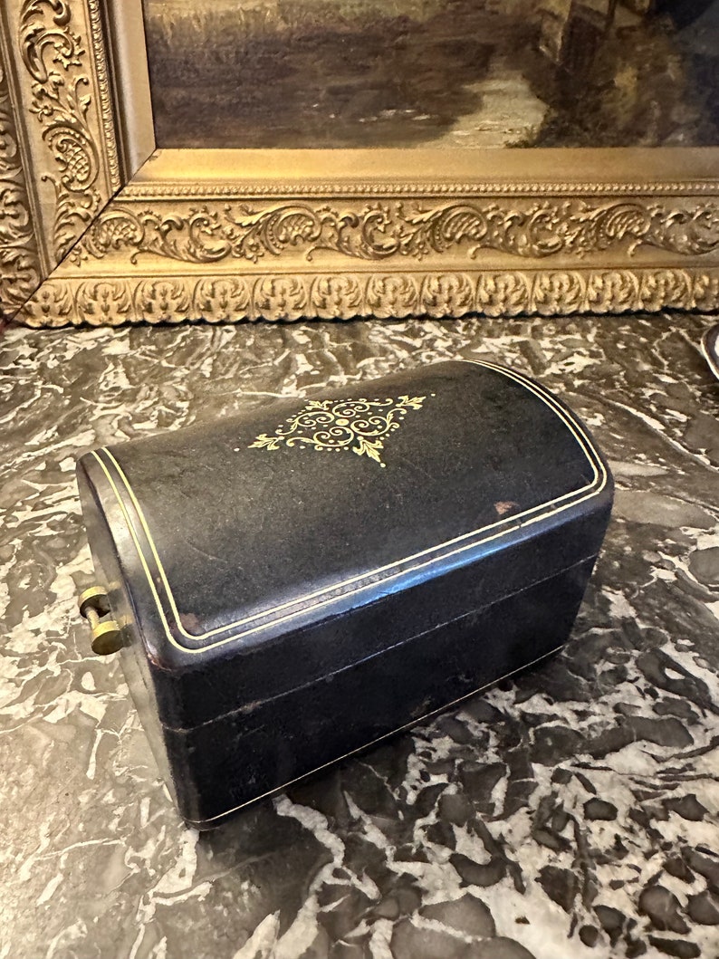 Puede incluir: Una caja de joyer&iacute;a negra, rectangular, con la parte superior curvada. La caja tiene ribetes dorados y un dise&ntilde;o decorativo dorado en la tapa. Se ve un cierre dorado en el lateral. La caja est&aacute; sobre una superficie de m&aacute;rmol.
