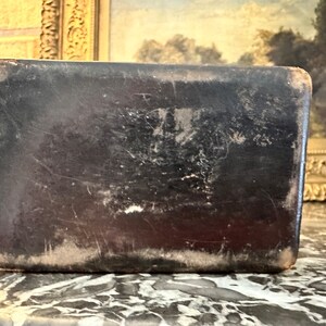 Puede incluir: Caja rectangular de cuero negro antiguo con un aspecto ligeramente desgastado. La caja presenta desgaste visible, con zonas m&aacute;s claras en los bordes y ara&ntilde;azos en la superficie, lo que sugiere edad y uso.