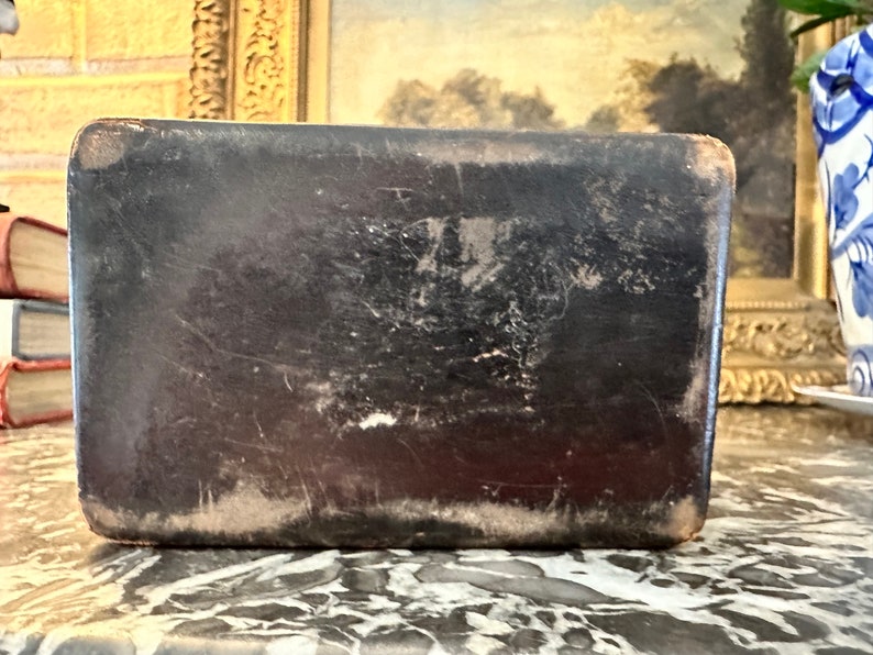 Puede incluir: Caja rectangular de cuero negro antiguo con un aspecto ligeramente desgastado. La caja presenta desgaste visible, con zonas m&aacute;s claras en los bordes y ara&ntilde;azos en la superficie, lo que sugiere edad y uso.