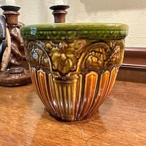 May include: A brown and green ceramic planter with an ornate design. The planter has a wide base and a narrow top, and it is decorated with raised leaves and flowers.