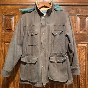 May include: A brown jacket with a green hood and multiple pockets. The jacket has a button-up front and a drawstring waist. The pockets are trimmed with green fabric. The jacket is hanging on a wooden hanger.