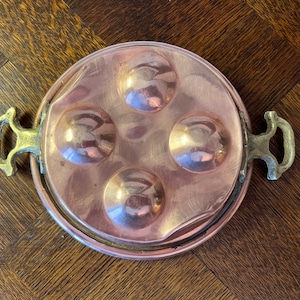 May include: A copper cooking pan with four round indentations and two brass handles. The pan is designed for cooking individual portions of food.