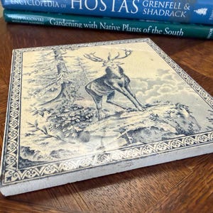 May include: A square ceramic tile featuring a blue and white illustration of a stag standing on a rocky outcrop near a forest. The tile has a decorative border pattern and a glossy finish, showcasing a detailed nature scene in a monochromatic style.