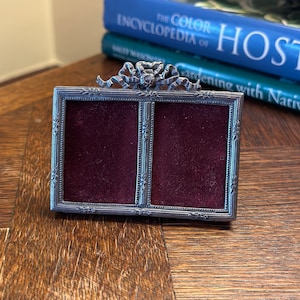 May include: A silver-toned picture frame with a velvet interior. The frame has a decorative bow at the top and a design of flowers and leaves around the edges.