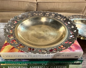 May include: Two brass serving dishes with intricate, ornate, cutout designs around the rim. The dishes are stacked on top of each other on a stack of books.