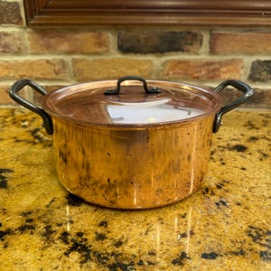May include: A round copper pot with a matching lid and two dark metal side handles. The pot features a polished metallic finish with visible signs of natural patina and wear, showcasing a warm, reflective surface and a sturdy, functional design.
