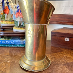 May include: A tall, polished brass boot jack with a wide, flared top and a round base. The boot jack has a small, circular design on the side.