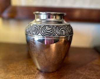 May include: A silver vase with a decorative, embossed design around the middle. The vase has a smooth, polished finish.