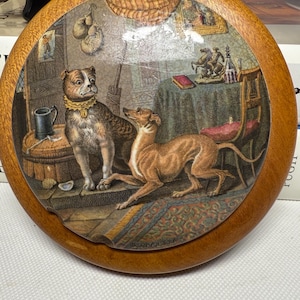 May include: Round wooden decorative piece featuring a detailed illustration of two dogs in a room. The dogs are a brown and white bulldog and a tan greyhound. The illustration is framed by a light brown wooden border.