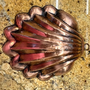 May include: A vintage copper shell-shaped baking mold with a fluted design. The mold has a small loop handle on the side.