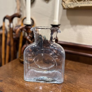 May include: A clear glass pitcher with a textured surface and a round indentation on the front. The pitcher has a wide mouth and a short spout.