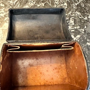 Puede incluir: Un estuche rectangular de cuero abierto con tapa abisagrada. El exterior es negro, mientras que el interior es de color marr&oacute;n claro. El estuche tiene un dise&ntilde;o simple y cl&aacute;sico, lo que sugiere que puede ser vintage o antiguo. El cuero muestra signos de desgaste.