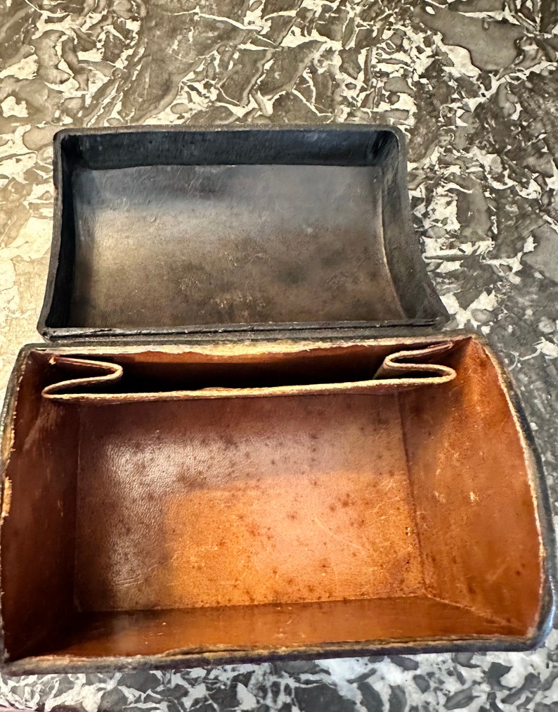 Puede incluir: Un estuche rectangular de cuero abierto con tapa abisagrada. El exterior es negro, mientras que el interior es de color marr&oacute;n claro. El estuche tiene un dise&ntilde;o simple y cl&aacute;sico, lo que sugiere que puede ser vintage o antiguo. El cuero muestra signos de desgaste.
