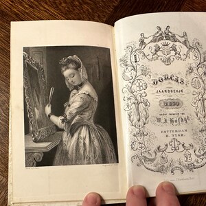 May include: An open book with a black and white illustration of a woman looking in a mirror on the left page. The right page features ornate text and decorative elements, including the words "DORCAS JAARBOEKJE 1854". The book is on a wooden surface.