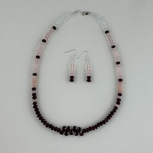 May include: A necklace and earring set featuring a combination of pink and dark red beads. The necklace has a strand of pink beads with dark red accents and a cluster of dark red beads at the center. The earrings mirror the necklace design.